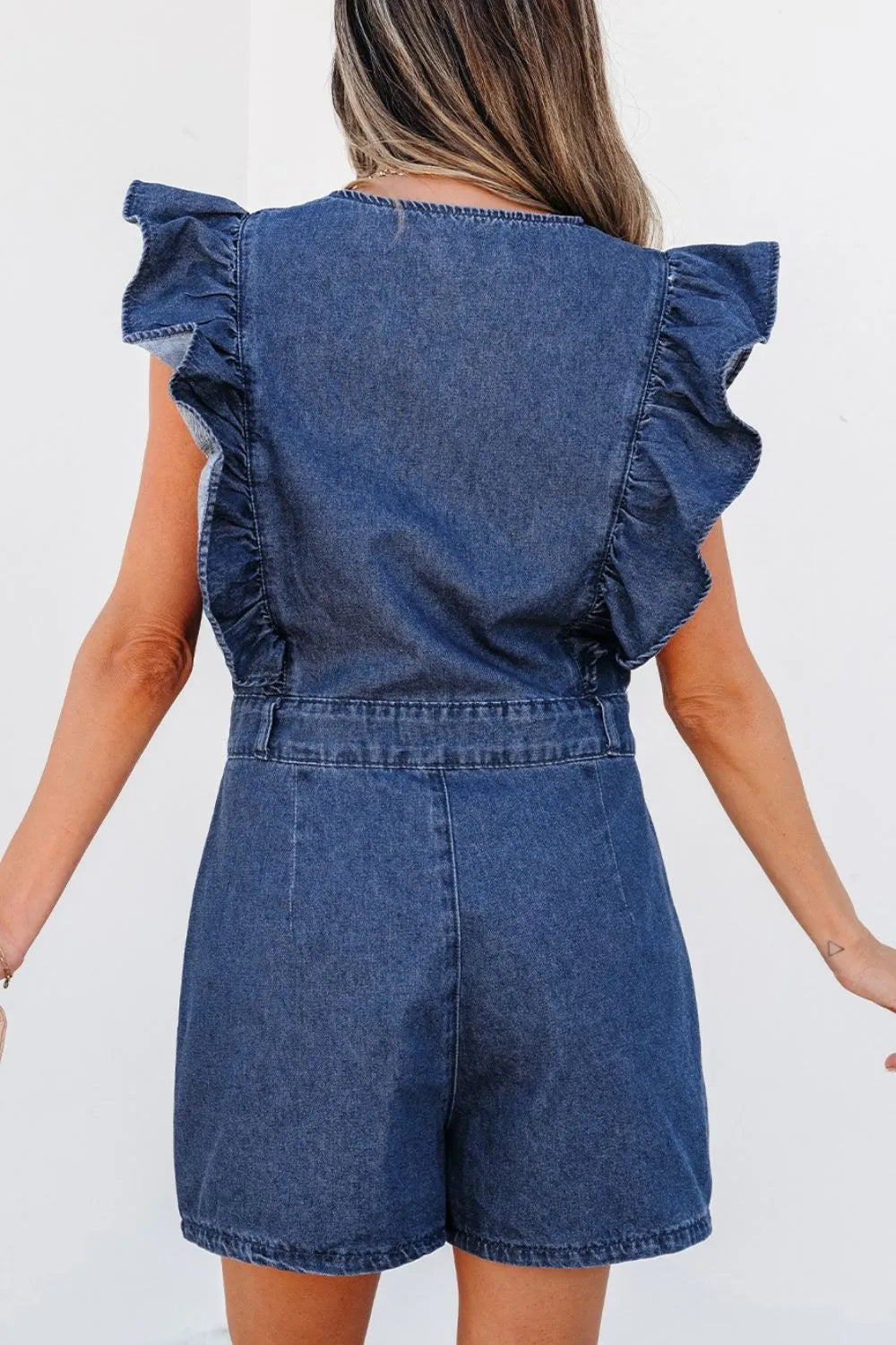 Woman seen from the back wearing a denim romper with ruffle sleeves, standing against a plain white background.