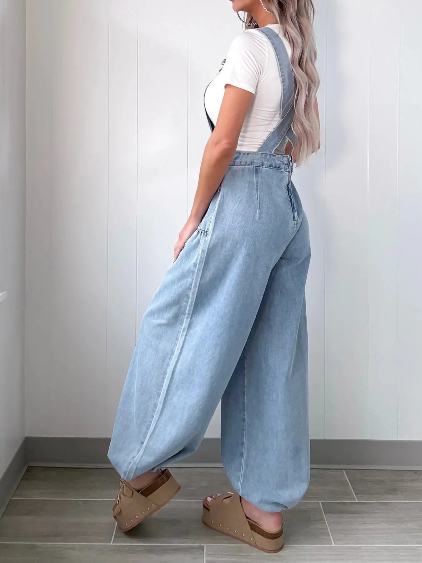 Side view of a woman in light blue denim wide-leg overalls with adjustable shoulder straps, paired with a white short-sleeve top and platform sandals, standing indoors.