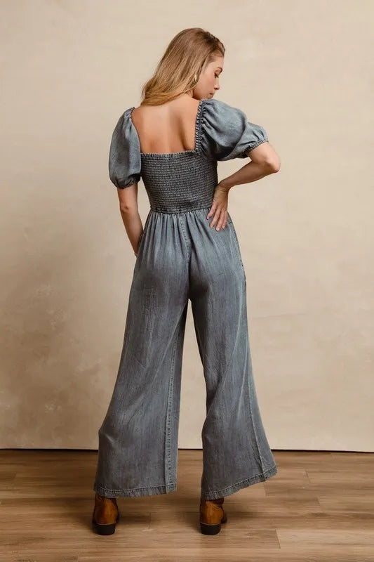 Back view of a woman wearing a vintage-style washed denim jumpsuit with a smocked bodice, puff sleeves, and wide-leg pants, paired with brown ankle boots
