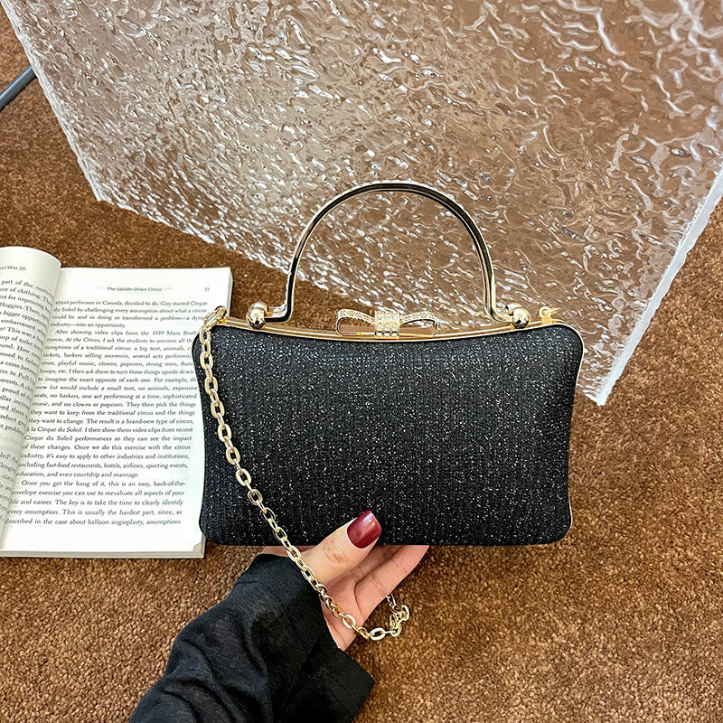 Black glittery handbag with gold chain held by a hand on a textured surface next to an open book.