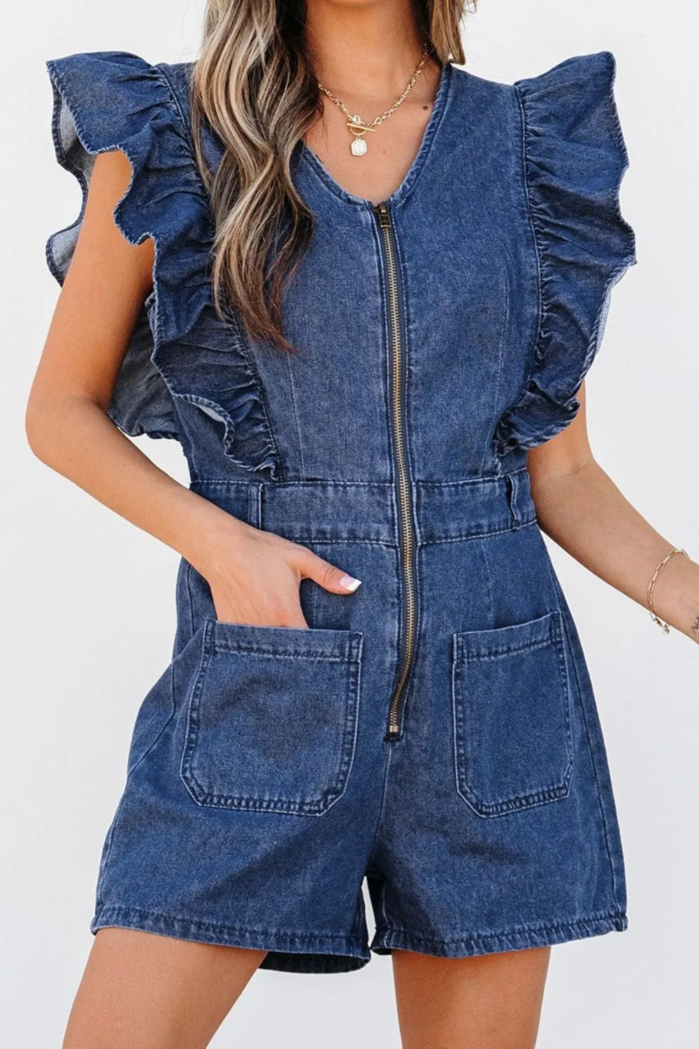 Woman wearing a blue denim romper with ruffle sleeves, front zipper, and large front pockets, accessorized with gold jewelry, standing against a plain white background.