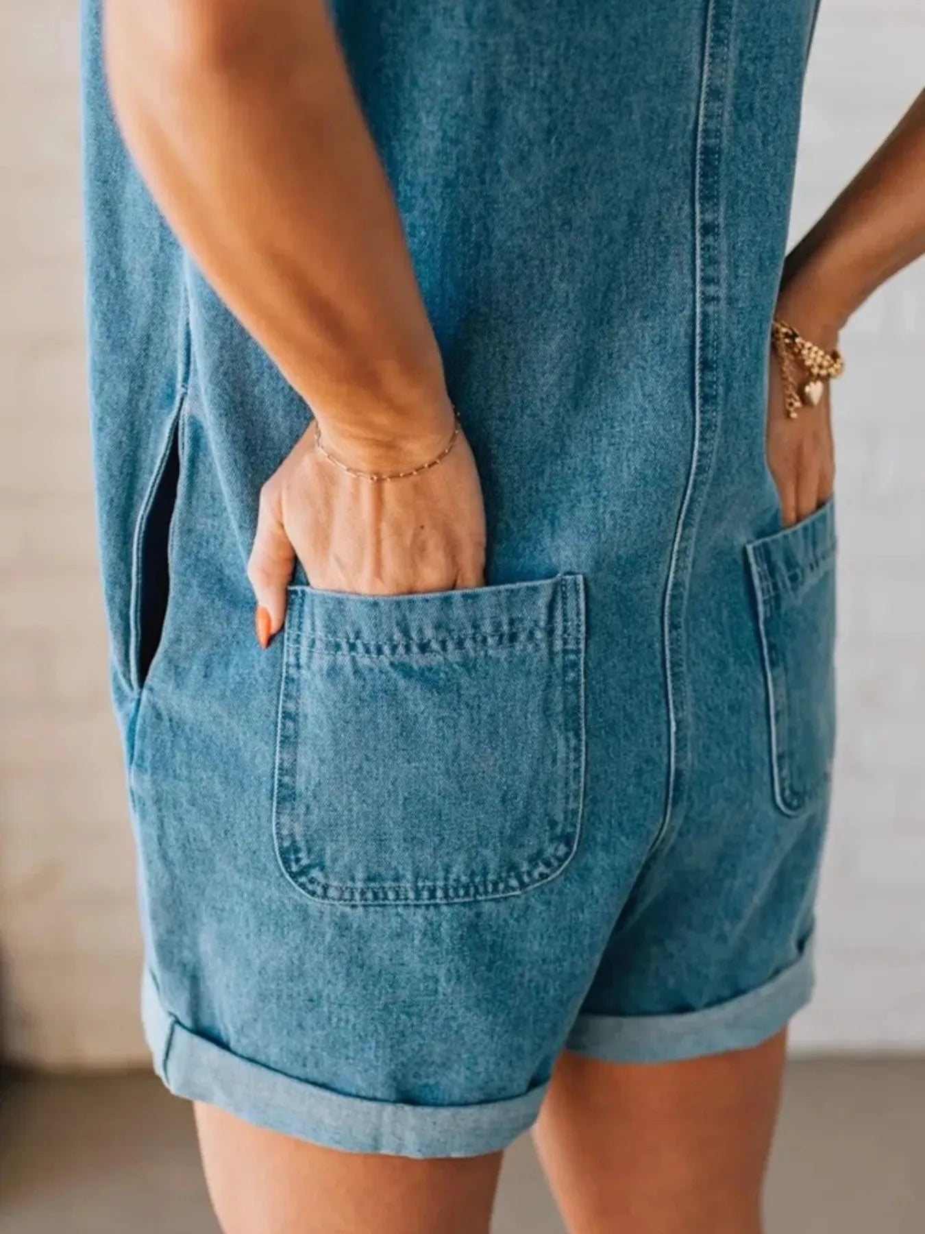 Close-up view of the back and side of a person wearing a light blue sleeveless denim romper, showing both hands placed in the back pockets. The person is wearing a thin bracelet and the shorts are rolled up at the hems.
