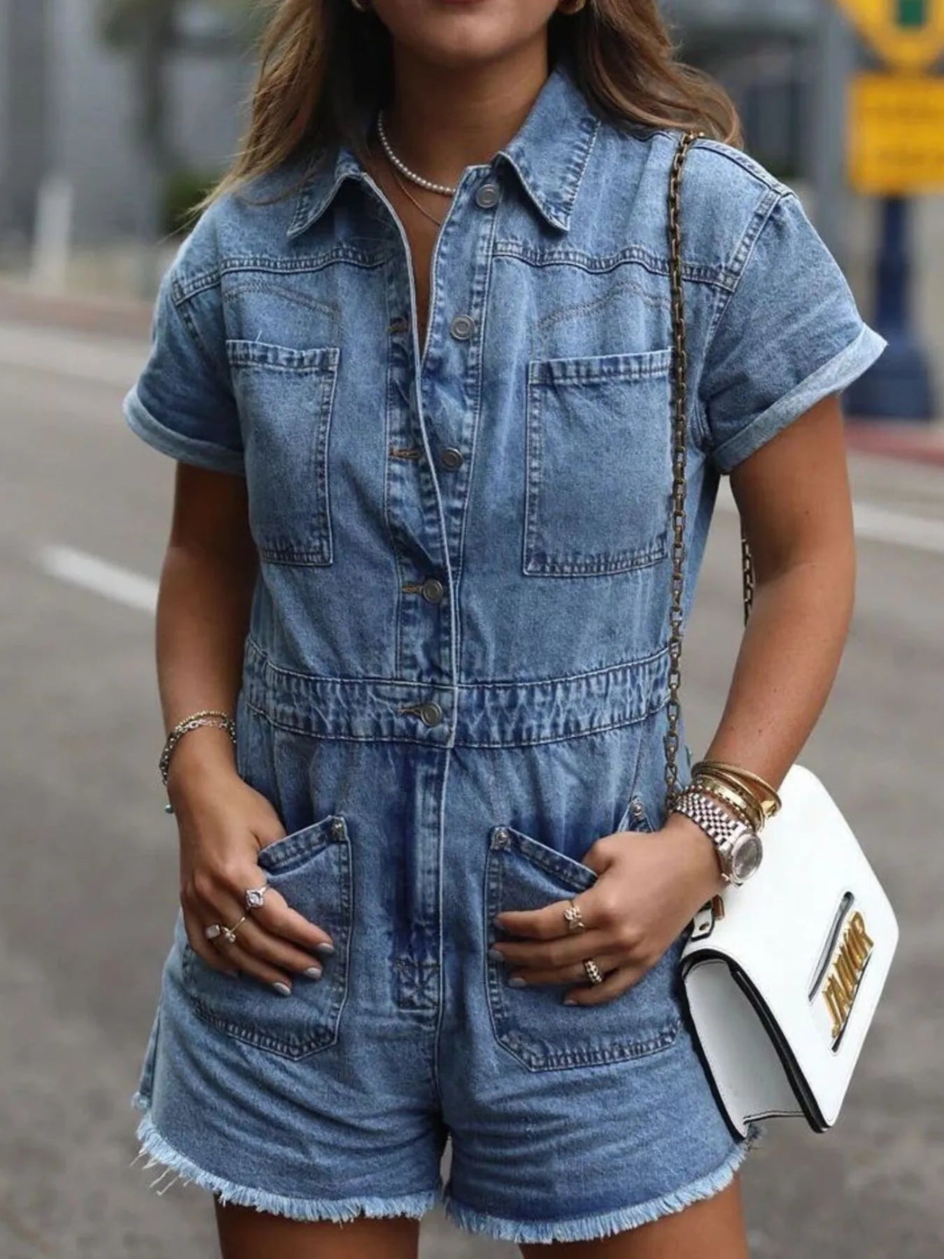 Woman wearing a short-sleeve denim romper with front pockets, accessorized with layered necklaces, rings, bracelets, a watch, and carrying a white purse with a gold chain strap, standing outdoors on a city street.