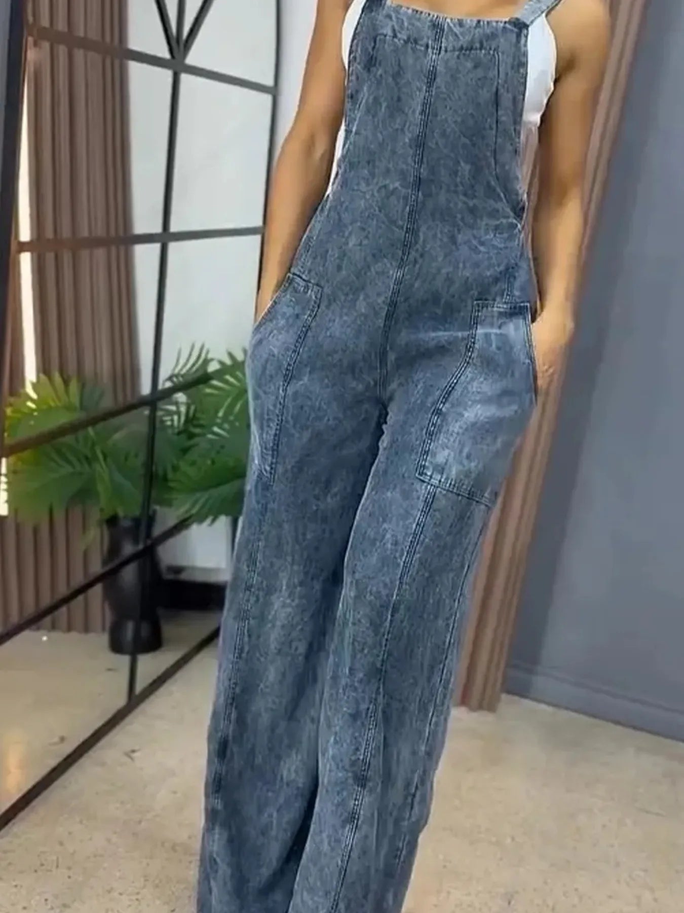 Closer view of a woman in stonewashed blue denim wide-leg overalls with front pockets, paired with a white tank top, standing with hands in her pockets indoors.