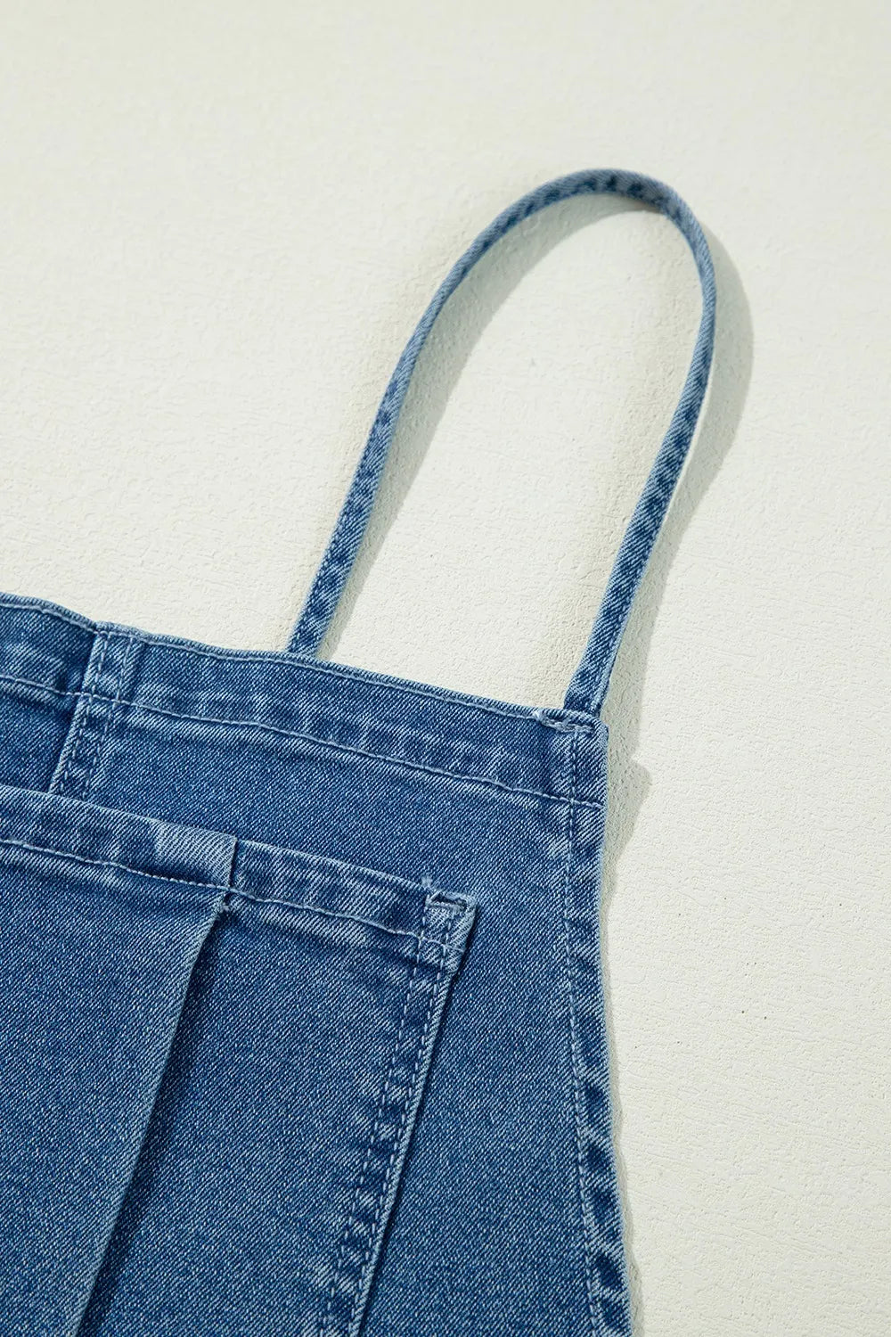 Close-up of the top portion of blue denim overalls, showing a thin shoulder strap and part of a front patch pocket, laid flat on a light-colored surface.