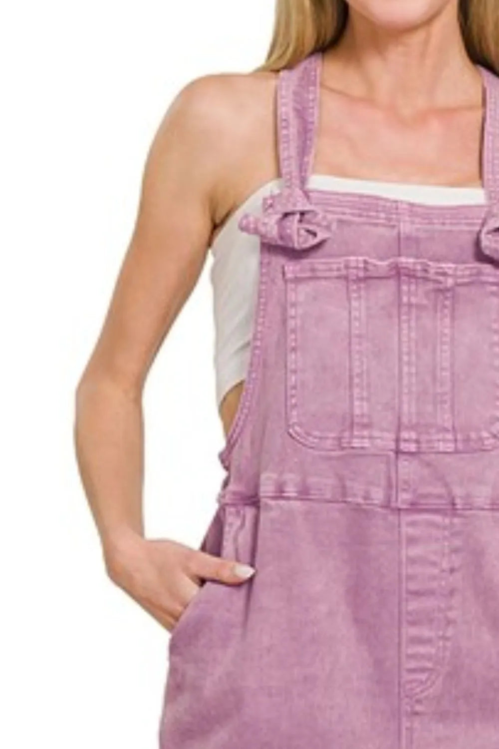 Woman wearing pastel purple denim overalls with front pockets, styled over a white tank top, standing with one hand in her pocket against a plain background.