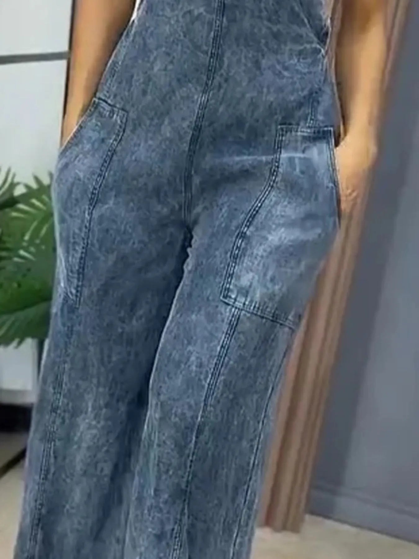 Close-up of the stonewashed blue denim wide-leg overalls, highlighting the pocket and seam details as the woman stands indoors with her hands in her pockets.