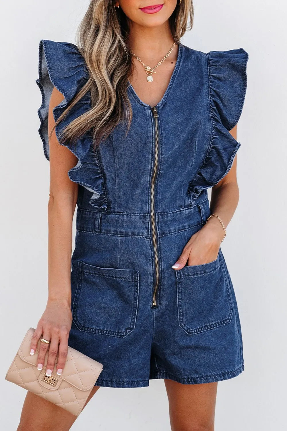 Woman wearing a denim romper with ruffle sleeves, front zipper, and large front pockets, accessorized with gold jewelry and holding a beige clutch purse.