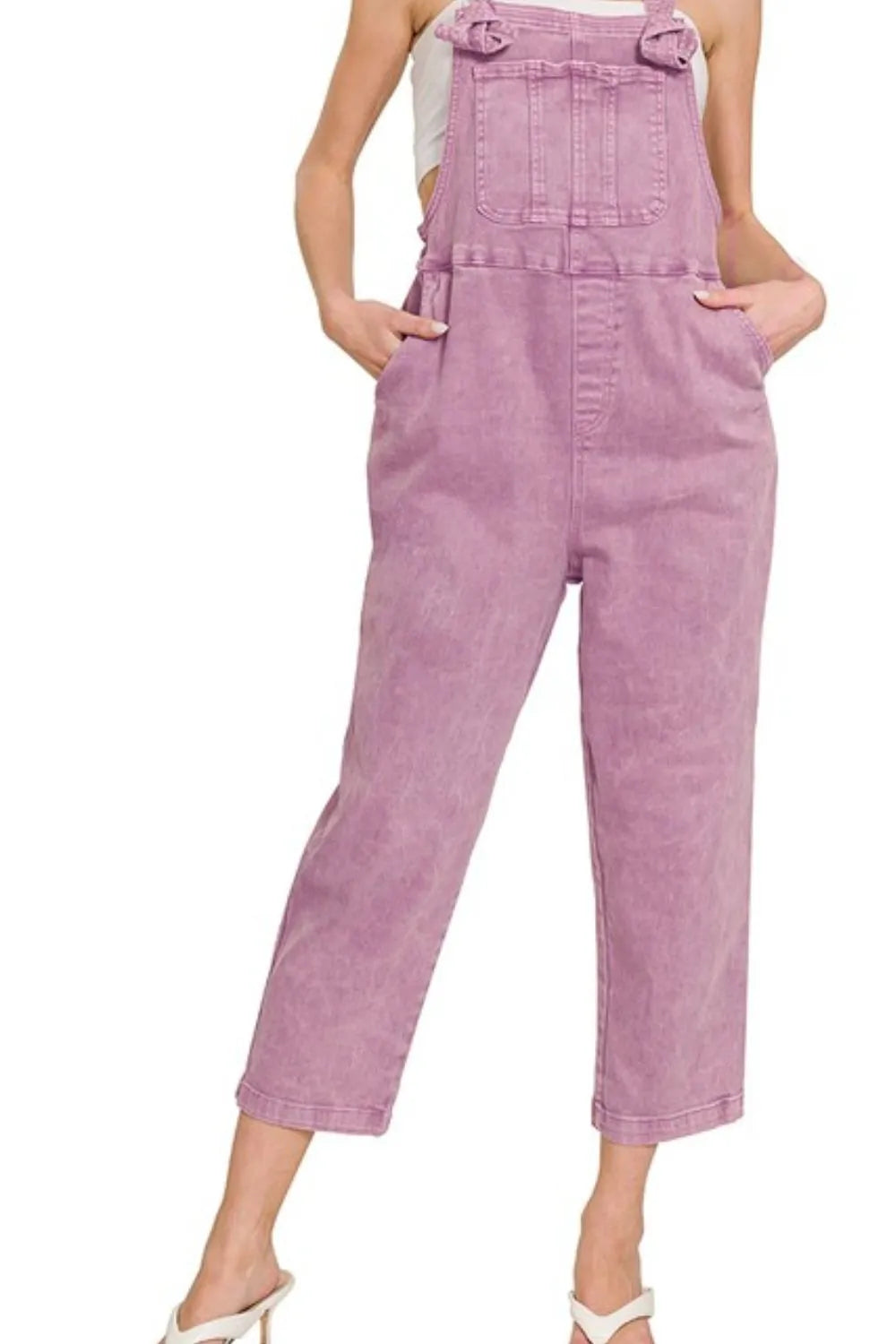 Woman wearing pastel purple denim overalls with front pockets, styled over a white top, standing with hands in pockets against a plain white background.