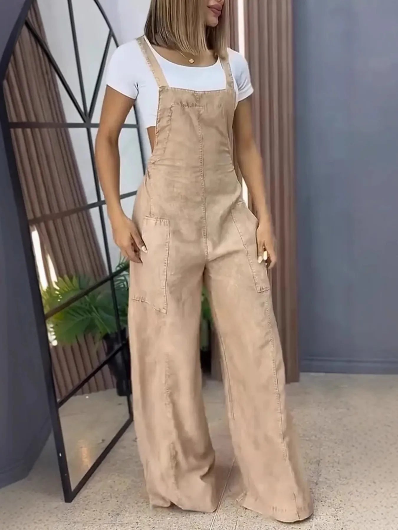 Woman wearing beige wide-leg overalls with front pockets, styled over a white short-sleeve top, standing indoors with hands relaxed at her sides.