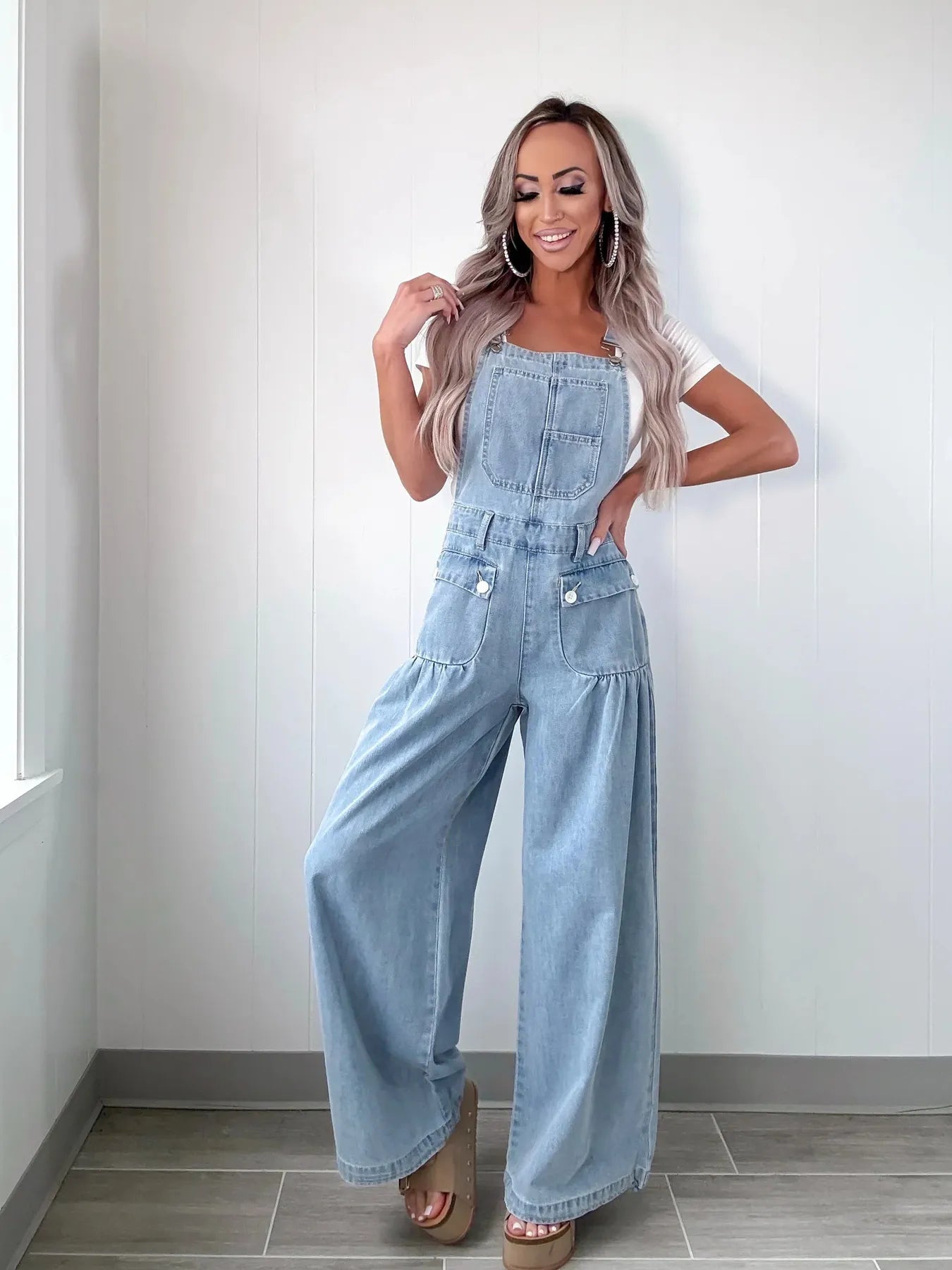 Woman posing in light blue denim overalls with a white T-shirt, wide-leg silhouette, and front pockets, standing against a plain white wall.