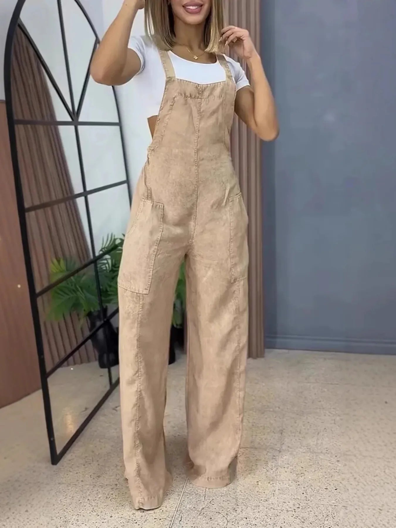 Woman posing in beige wide-leg overalls with large front pockets, paired with a white fitted top, holding one hand to her hair in a modern indoor setting.