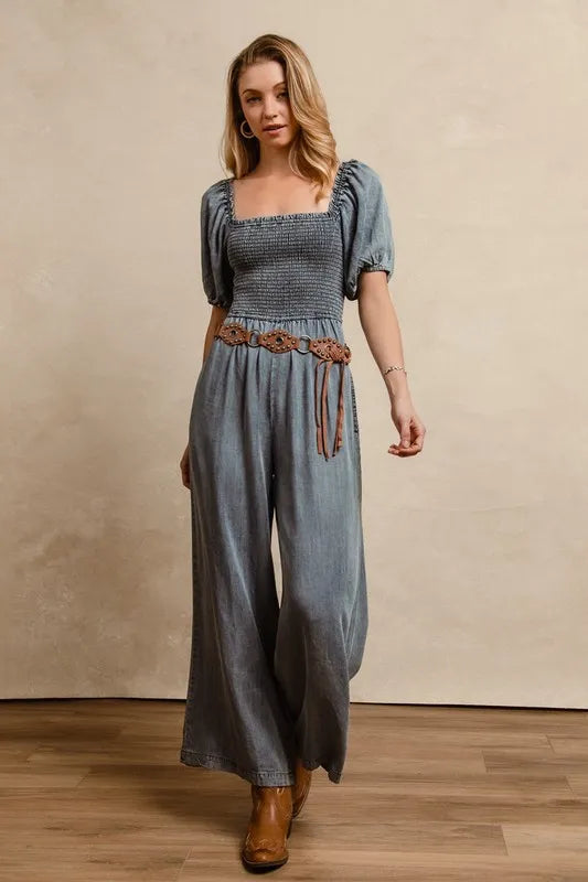 Woman wearing a vintage-inspired washed denim jumpsuit featuring a smocked bodice, puff sleeves, wide-leg pants, and a woven brown belt, styled with ankle boots
