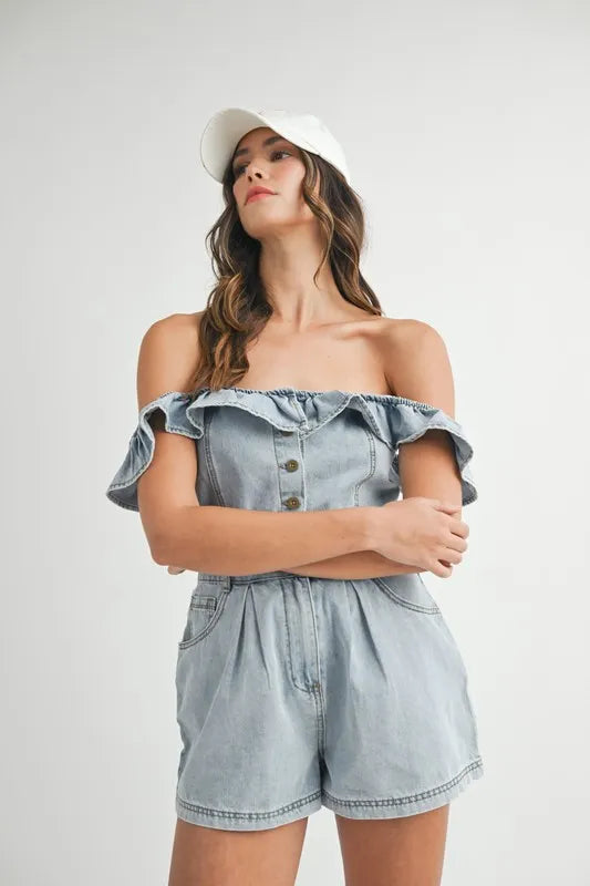 Woman wearing a light-washed, off-shoulder denim romper with ruffled neckline, front button detailing, and pleated shorts, styled with a white baseball cap.