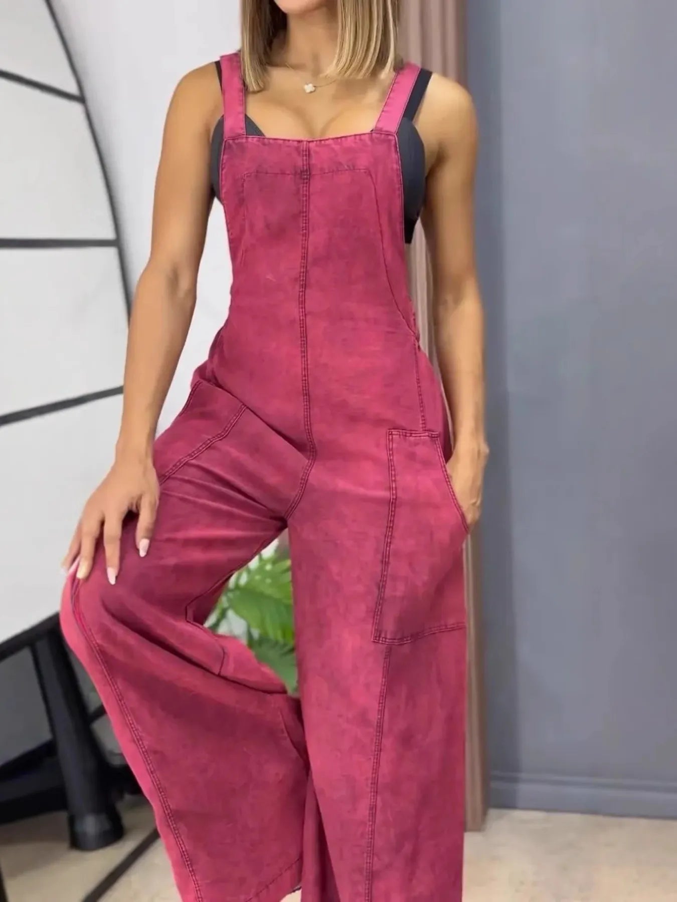 Woman wearing magenta wide-leg overalls with front patch pockets, styled over a black fitted top, standing indoors with one hand in the pocket.