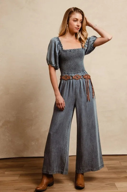 Woman posing in a vintage-inspired smocked denim jumpsuit with puff sleeves and ankle boots on a wooden floor background