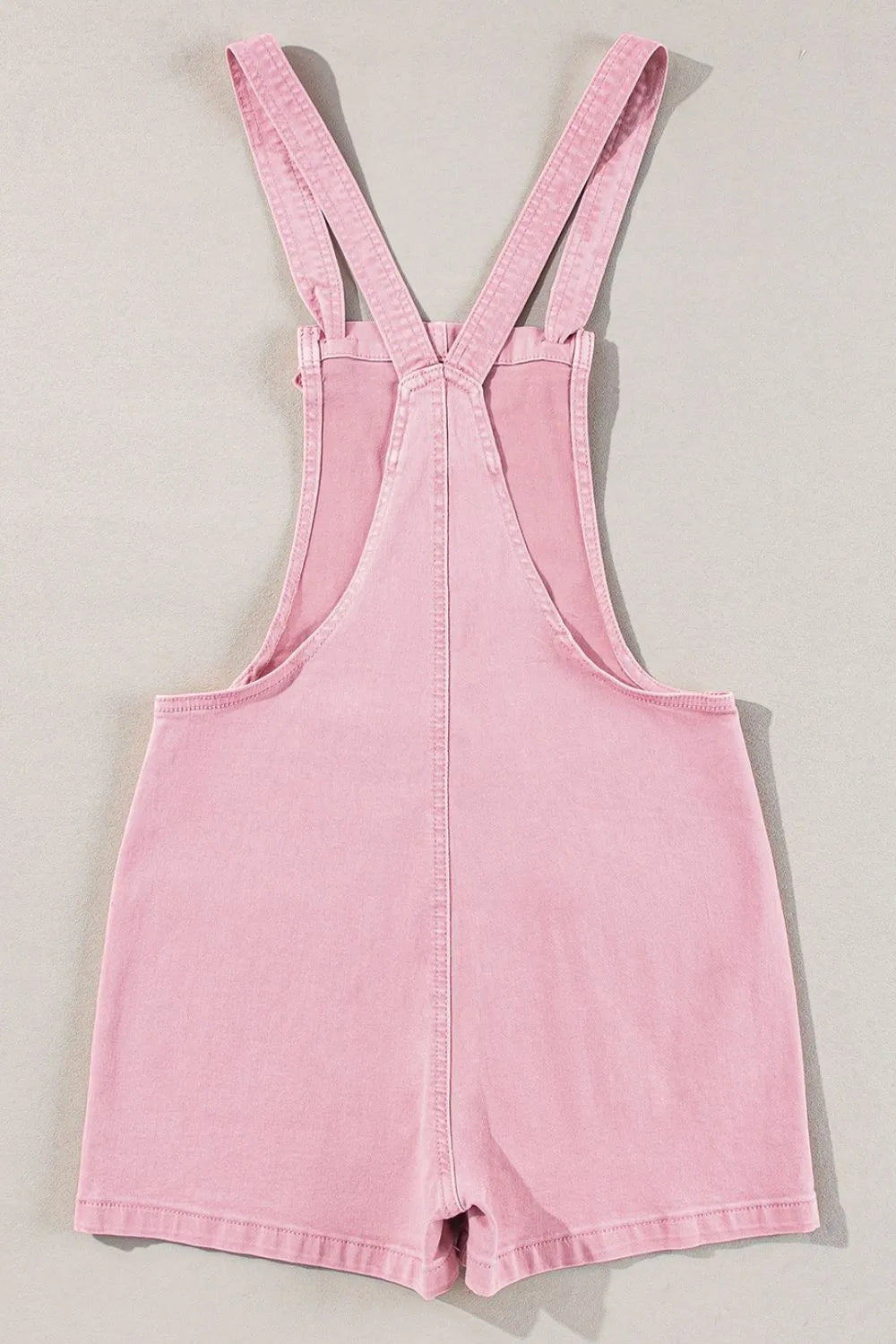 Light pink denim short overalls with crossed straps, laid flat.