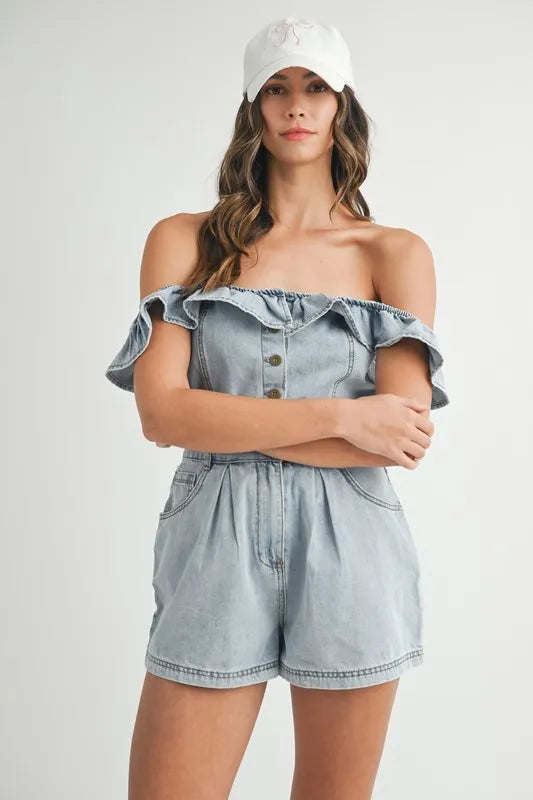 Front view of a woman wearing a light-wash, off-shoulder denim romper featuring a ruffled neckline, button-down front, and pleated shorts, styled with a white baseball cap.