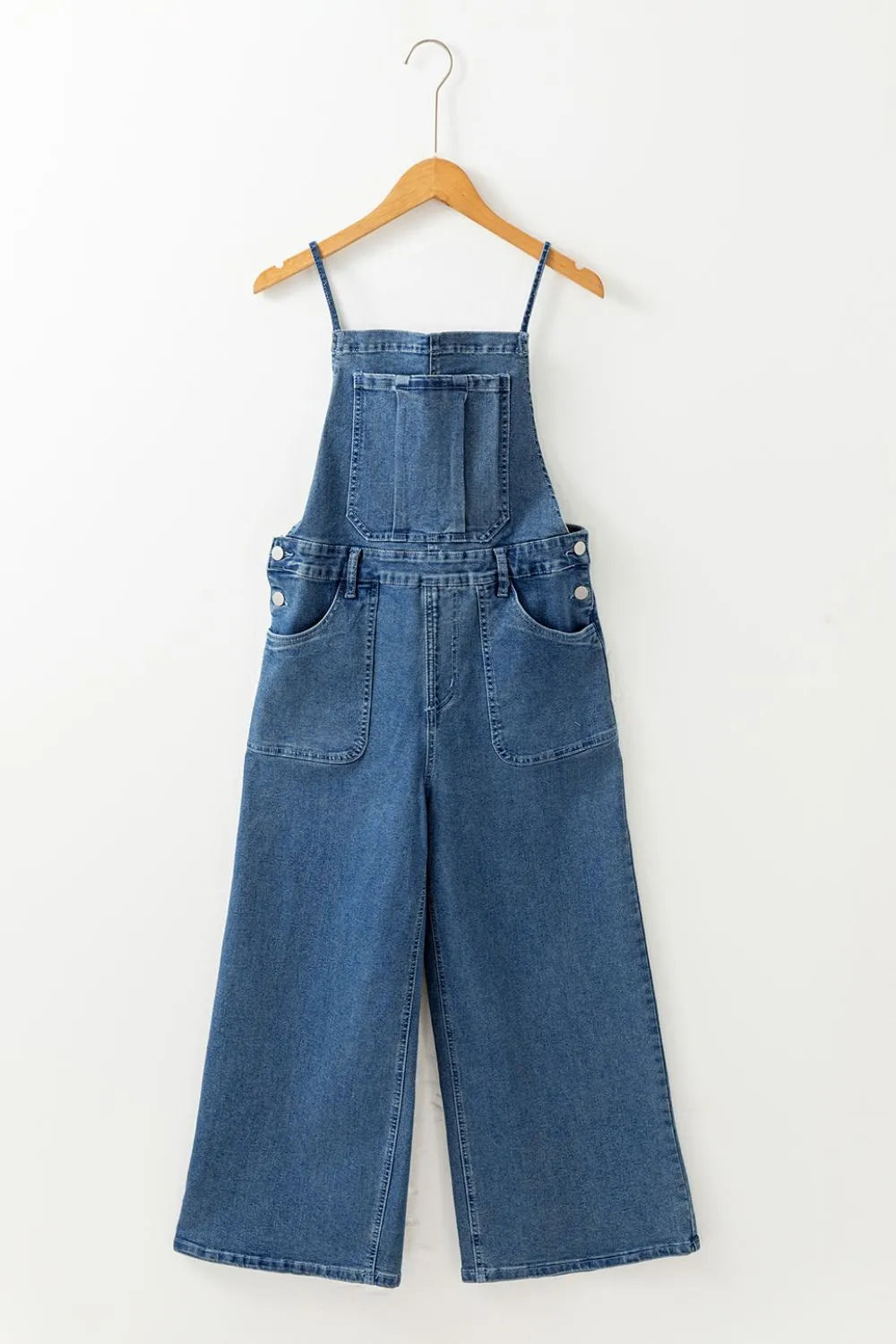 Blue denim overalls with wide legs, front patch pockets, a square bib with a chest pocket, and thin adjustable shoulder straps, displayed hanging on a wooden hanger against a white background