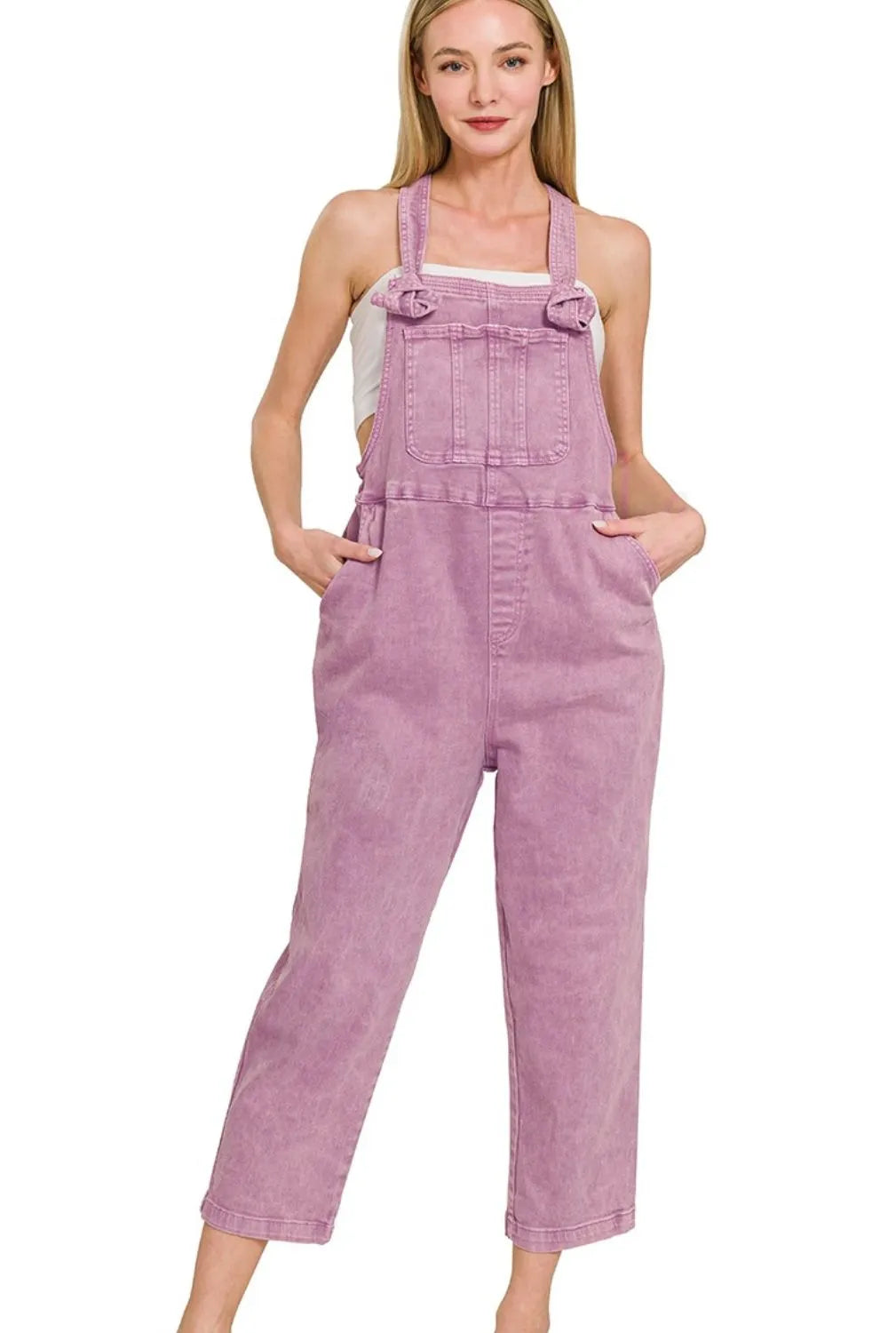 Woman wearing light purple denim overalls with front pockets, styled over a white top, standing with hands in pockets against a plain background