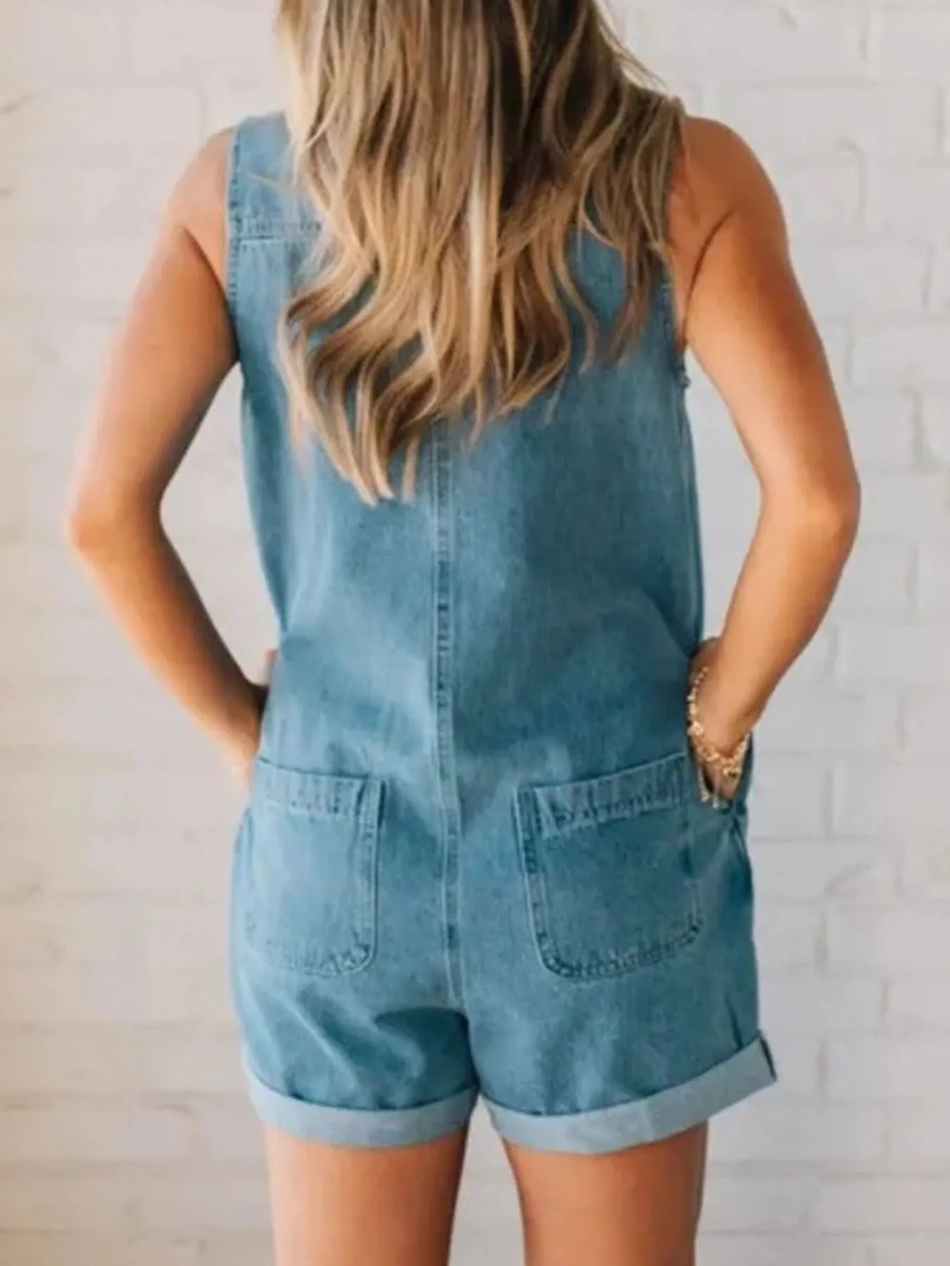 Rear view of a woman standing against a light brick wall, wearing a sleeveless blue denim romper with rolled-up shorts. Both hands are in her back pockets, and her long, wavy blonde hair falls over her shoulders.