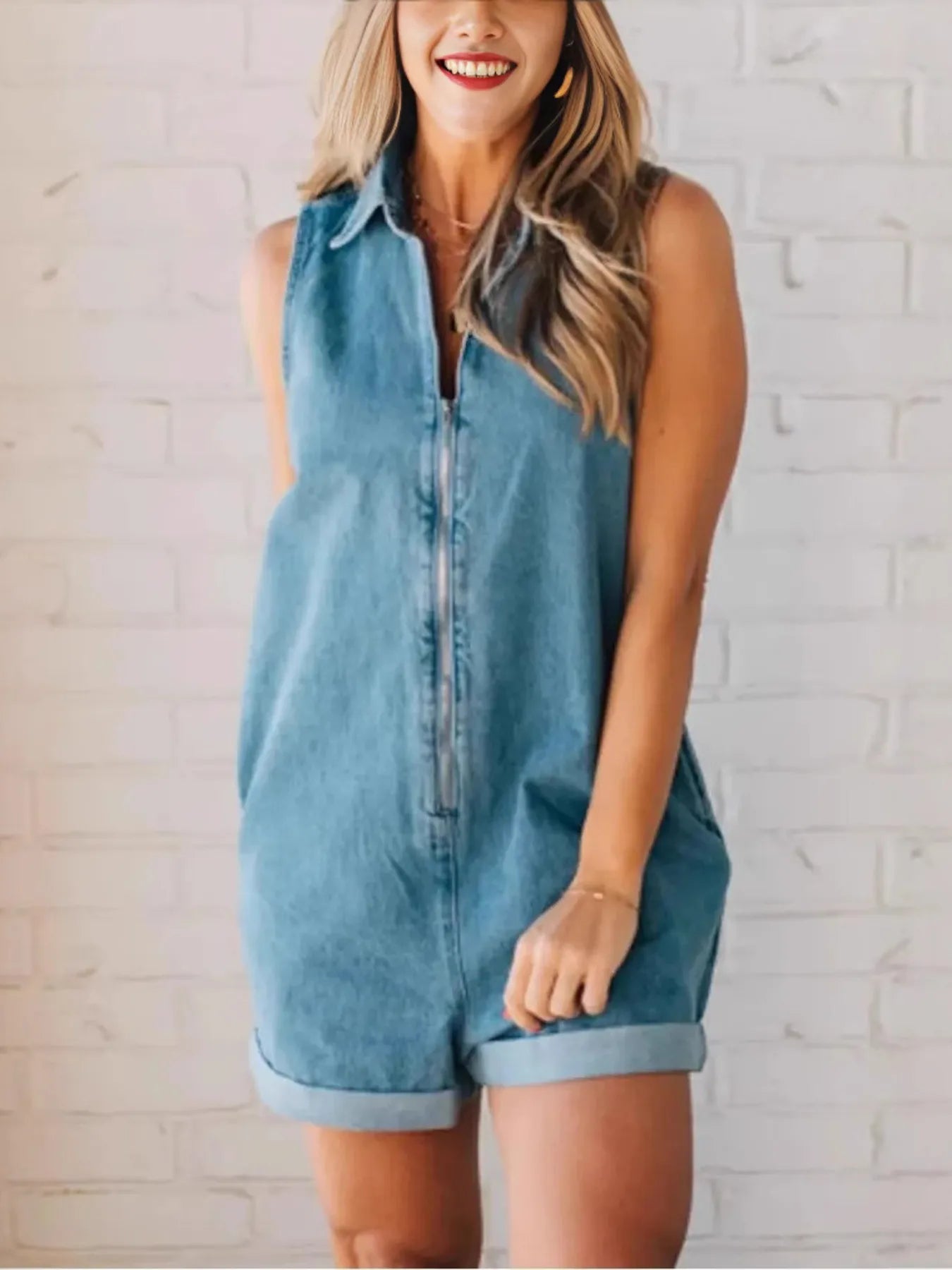 Woman modeling a loose-fitting, sleeveless denim romper with a collar and rolled cuffed shorts, standing with one hand in her pocket in front of a white brick wall.