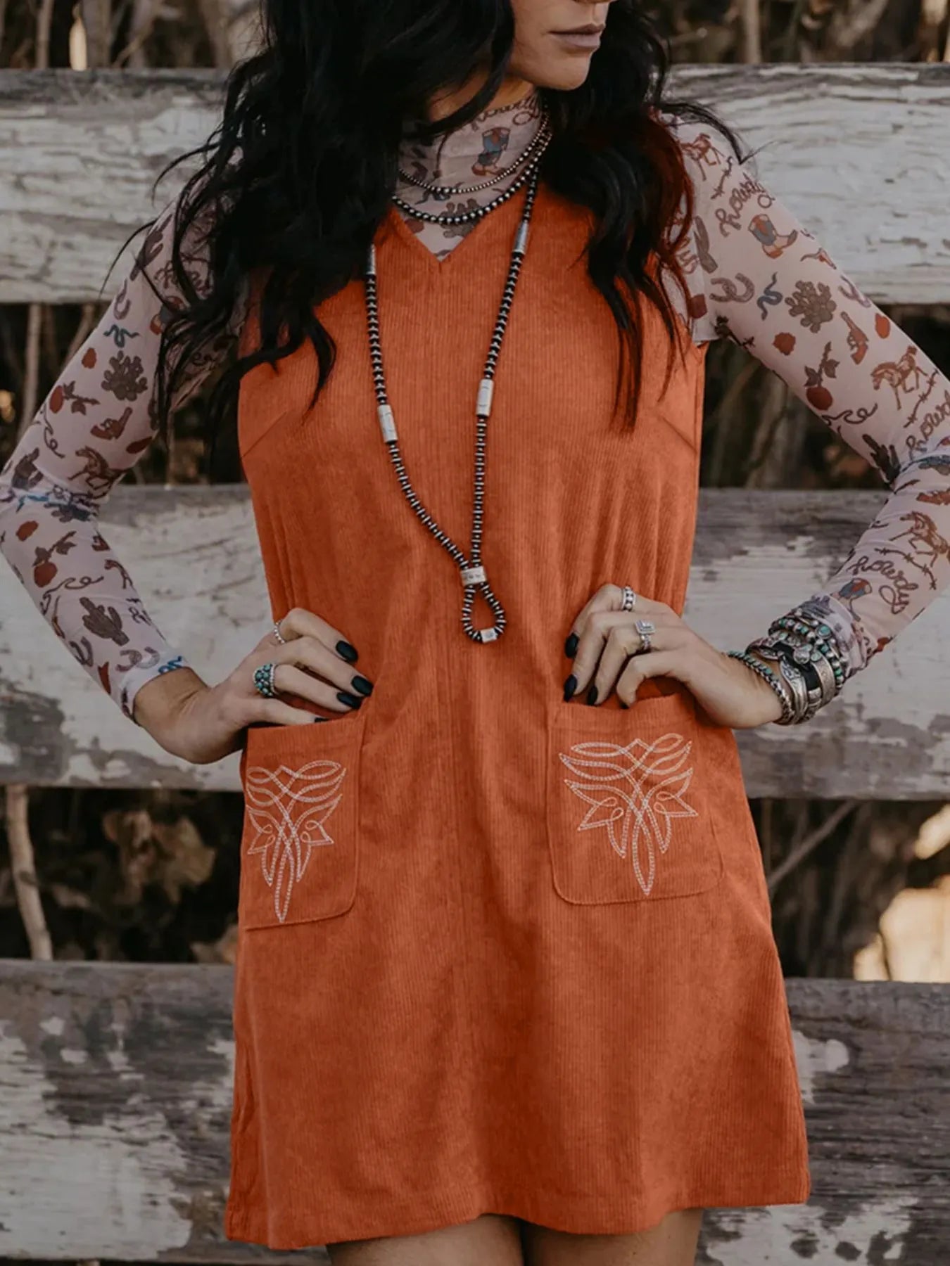 Model styling a sleeveless rust corduroy dress with hand embroidery on pockets and a sheer mesh cactus-printed top underneath, standing confidently with hands on waist – trendy bohemian outfit for women.