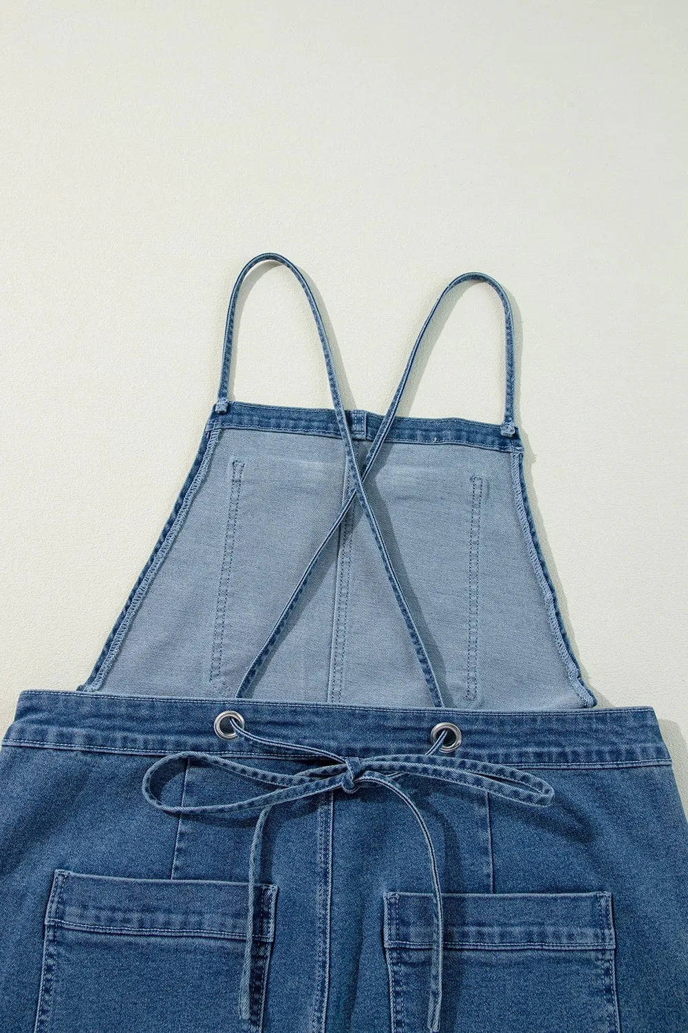 Close-up of blue denim overalls laid flat on a light surface, featuring a square bib, two thin shoulder straps crossing at the back, a drawstring tie at the waist, and large front patch pockets.