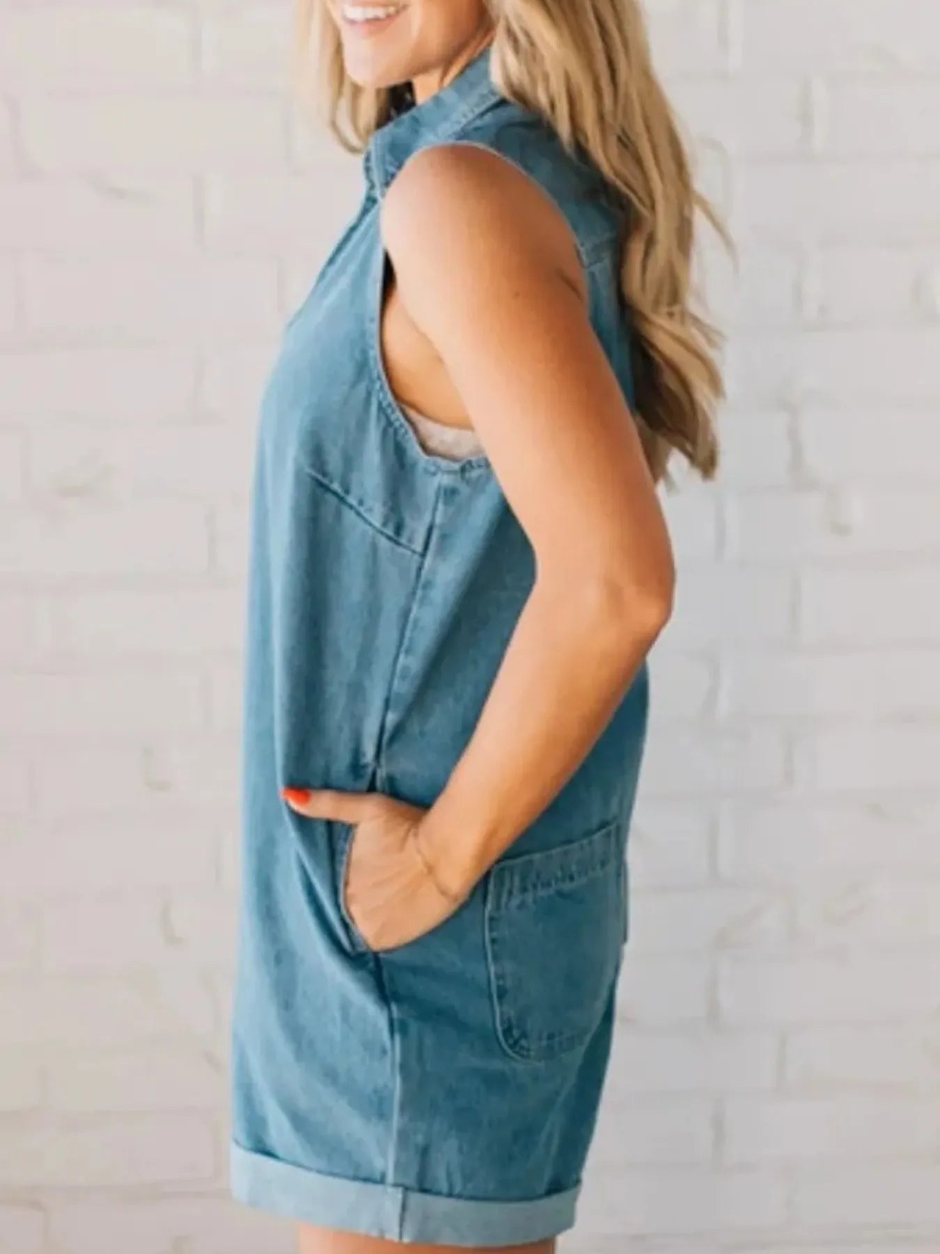 Side profile of a woman in a sleeveless denim romper with large side pocket, collar, and rolled shorts, with her hand in the pocket, standing by a white brick wall.