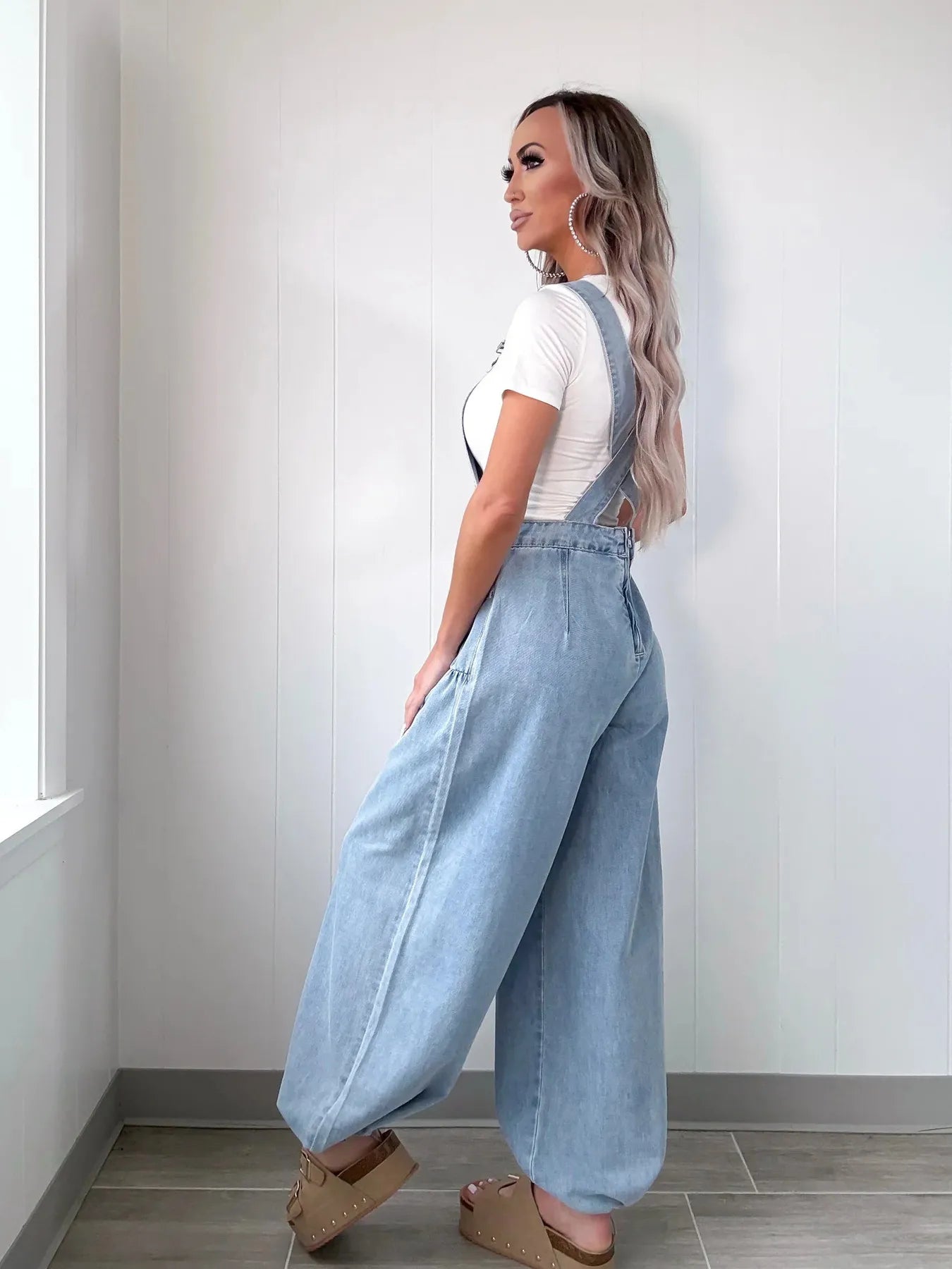 Side profile of a woman in light blue denim overalls, showing the loose wide-leg fit and crossover straps at the back, styled with tan platform sandals and a white short-sleeve top, indoors.