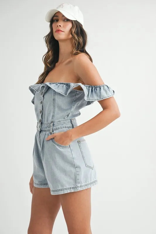 Side view of a woman in a light-washed off-shoulder denim romper featuring ruffled neckline, front buttons, and relaxed-fit shorts, styled with a white baseball cap and hands in pockets.