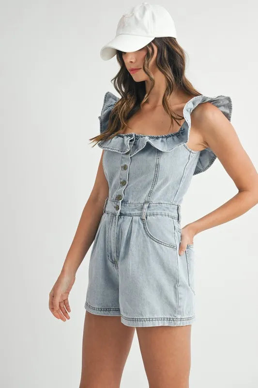 Woman wearing a light-washed denim romper with ruffled shoulder straps, a ruffled neckline, front button closure, and pleated shorts, styled with a white baseball cap.
