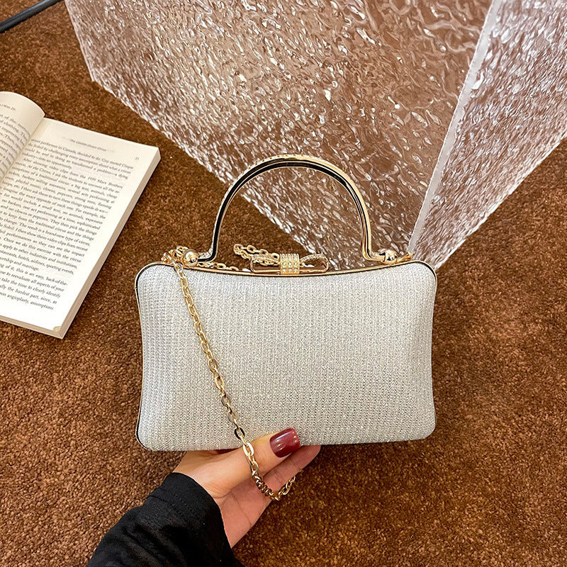 White handbag with gold chain and handle held by a hand on a brown surface next to an open book.