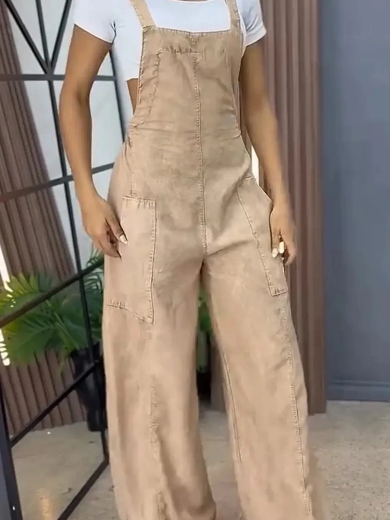 Woman in beige wide-leg overalls with front pockets, styled over a white short-sleeve top, standing with one hand in her pocket against a neutral indoor background.