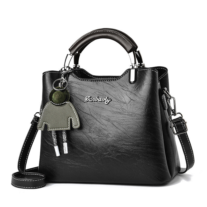 Front view of a stylish black faux leather women's handbag with a top handle, detachable shoulder strap, and cute green doll keychain accessory.