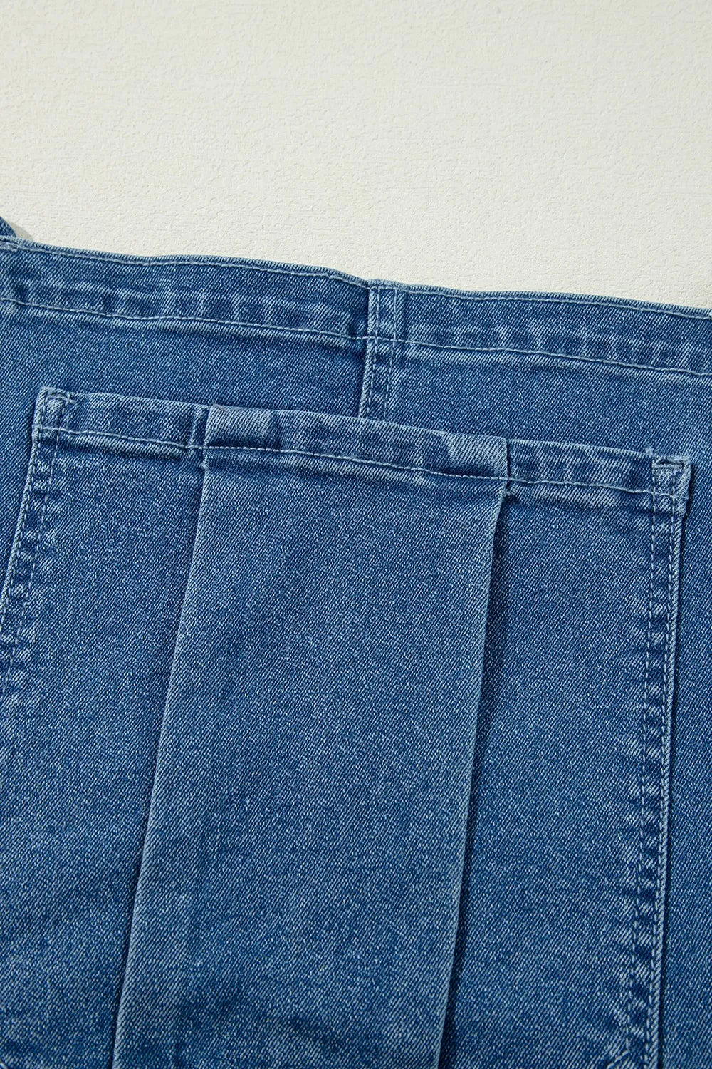 Close-up image of blue denim fabric showing a portion of a garment, likely jeans or overalls. The image focuses on a large rectangular pocket with a wide belt loop or strap sewn down the middle, set against a plain light background. The visible stitching and seams highlight the texture and construction of the denim material.