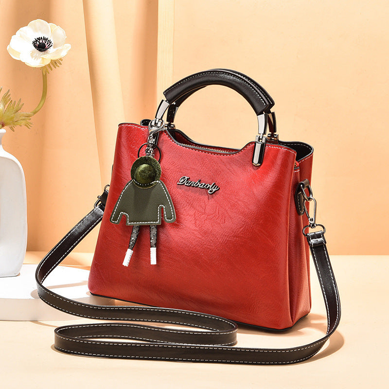 Bold red faux leather women's handbag with black top handle, adjustable strap, and cute green doll keychain, styled against a soft beige backdrop.