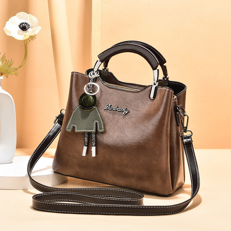 Chic brown faux leather women's handbag with top handle, adjustable shoulder strap, and green doll keychain accessory, styled against a soft beige background.