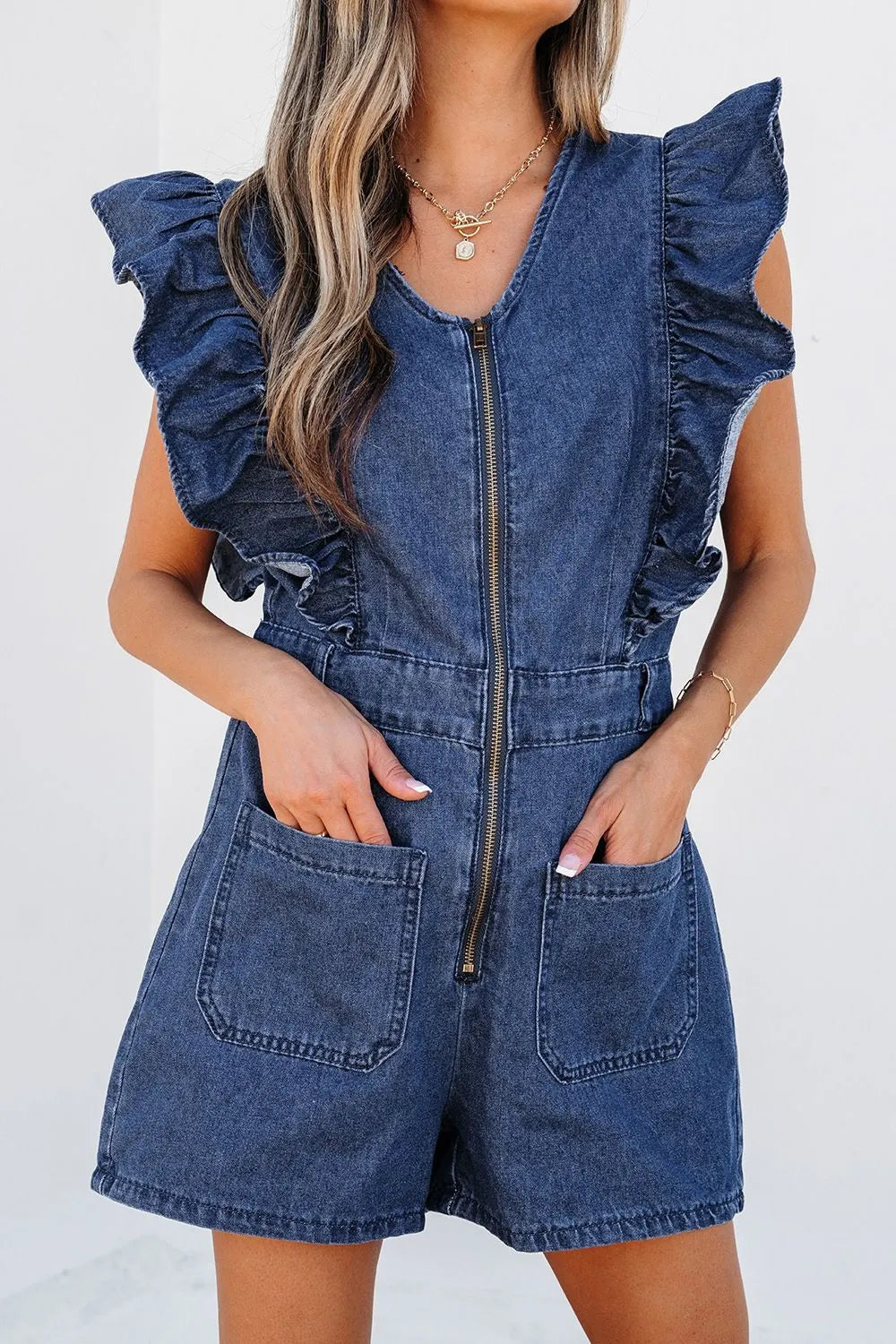 Woman wearing a denim romper with ruffle sleeves, front zipper, and large front pockets, accessorized with gold jewelry.
