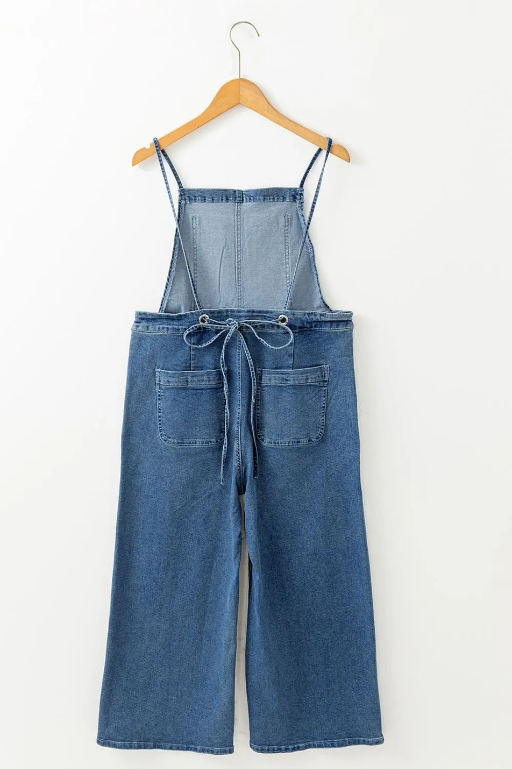 Blue denim overalls with wide legs, front patch pockets, a drawstring tie at the waist, and thin shoulder straps, displayed on a wooden hanger against a white background.