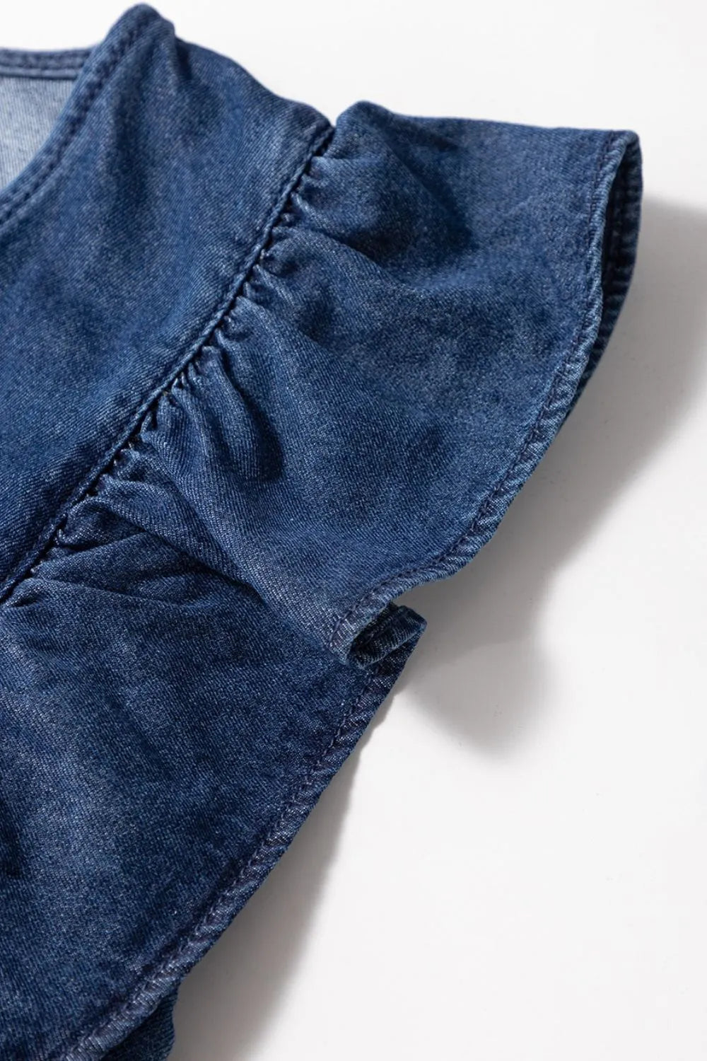 Close-up of a ruffle sleeve on a blue denim romper, showing the fabric texture and stitching detail against a white background.