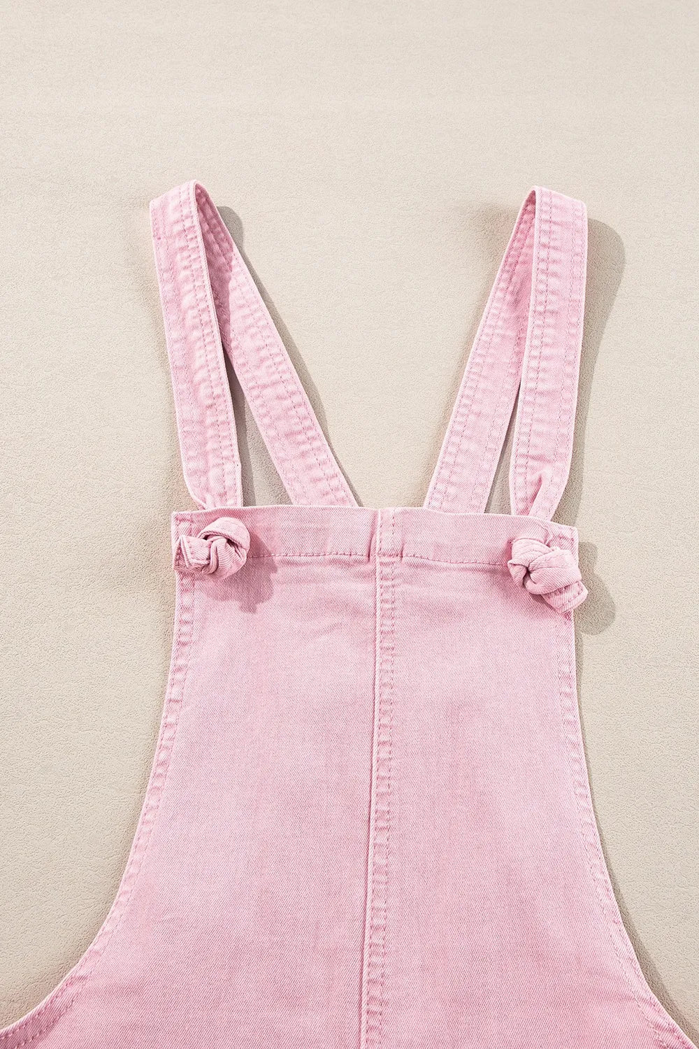 Pink denim overalls with knotted shoulder straps, laid flat.