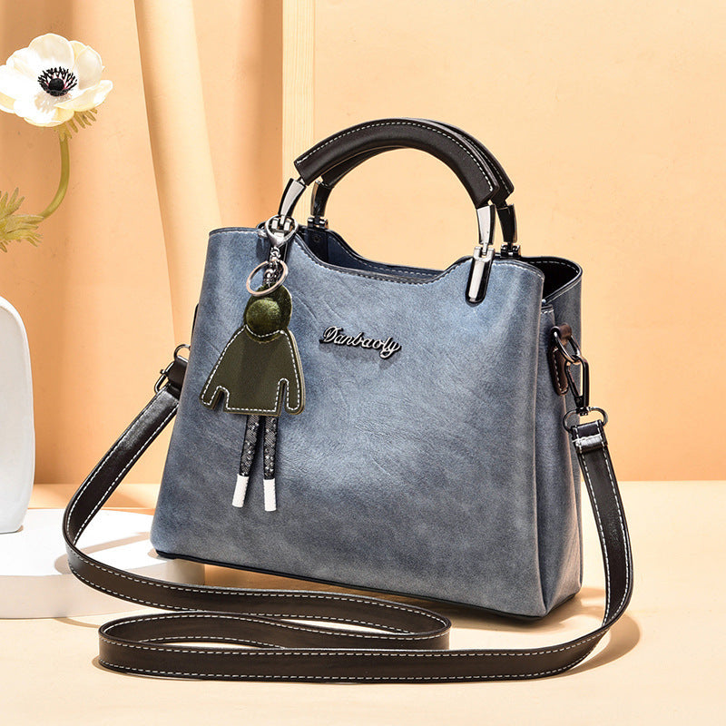 Stylish blue faux leather women's handbag with black top handle, long shoulder strap, and playful green doll keychain, displayed against a peach-toned backdrop.