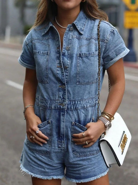 Woman wearing a short-sleeve denim romper with front pockets, accessorized with layered necklaces, rings, bracelets, a watch, and carrying a white purse with a gold chain strap, standing outdoors on a city street.