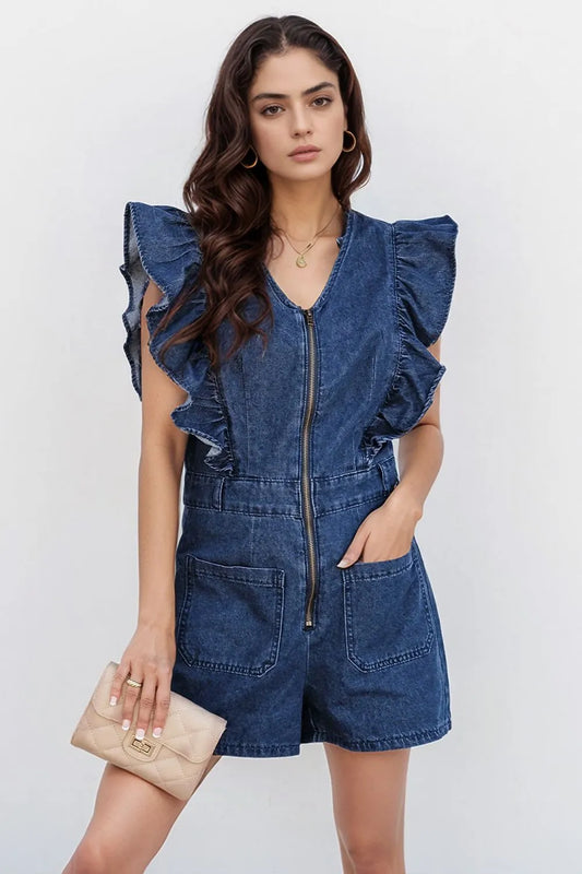 Woman wearing a denim romper with ruffle sleeves, front zipper, and large front pockets, accessorized with gold jewelry and holding a beige clutch purse.