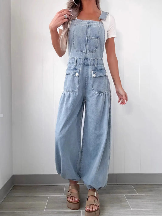 Woman wearing light blue denim overalls with wide legs and front pockets, styled over a white fitted short-sleeve top, standing in a minimal indoor setting