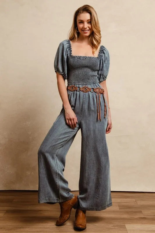 Woman wearing a washed denim smocked jumpsuit with puff sleeves and wide-leg fit, styled with a boho leather belt and ankle boots.