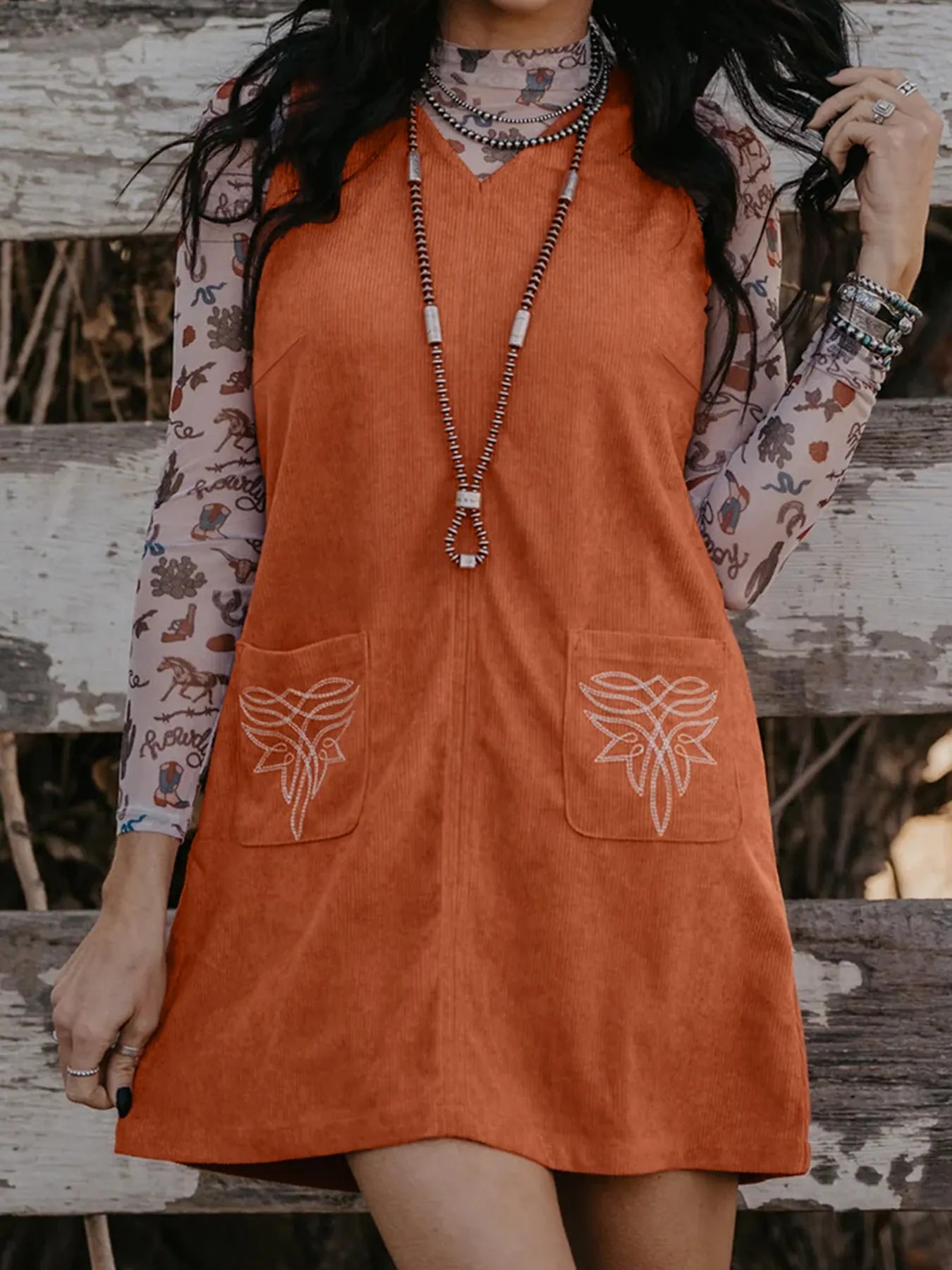 Front view of a woman wearing a rust-colored corduroy mini dress with V-neck, floral embroidery on front pockets, layered necklaces, and sheer cactus print inner top – perfect for boho fall fashion for women.