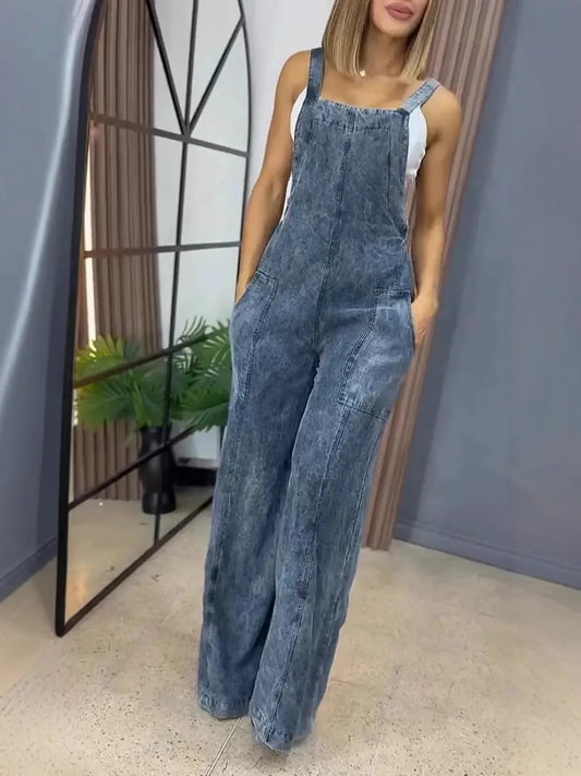 Woman wearing stonewashed blue denim wide-leg overalls with front pockets, styled over a white tank top, standing indoors with hands in her pockets.
