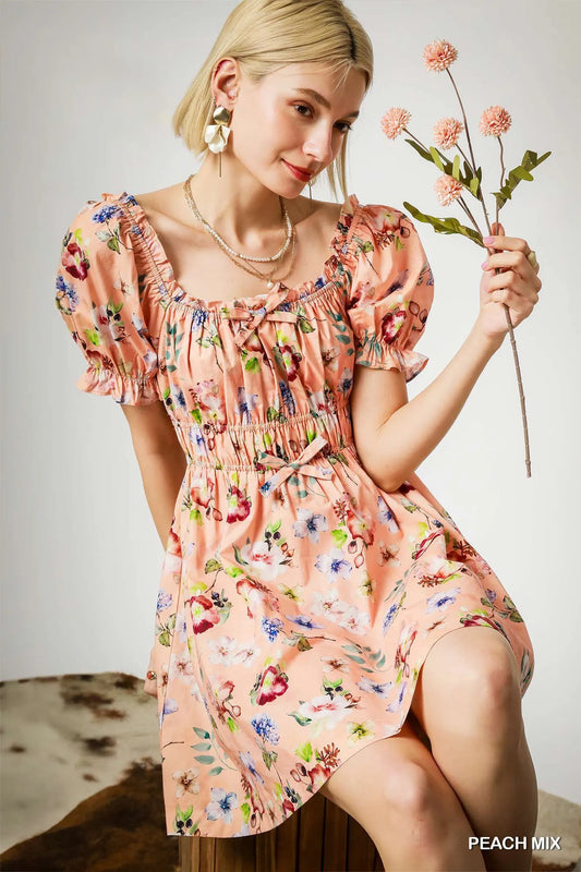 Woman wearing a peach floral ruched mini dress with puff sleeves, sitting and holding pink flowers